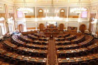Vista ampla de um plenário legislativo ornamentado, com cadeiras vazias e lustres