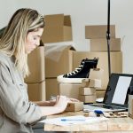 Microempreendedora embala caixas para sua loja online, cercada por materiais de envio e um laptop.