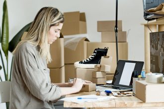 Microempreendedora embala caixas para sua loja online, cercada por materiais de envio e um laptop.