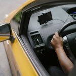 Vista superior de uma pessoa dirigindo um táxi amarelo equipado com navegador GPS em uma rua urbana durante o dia.