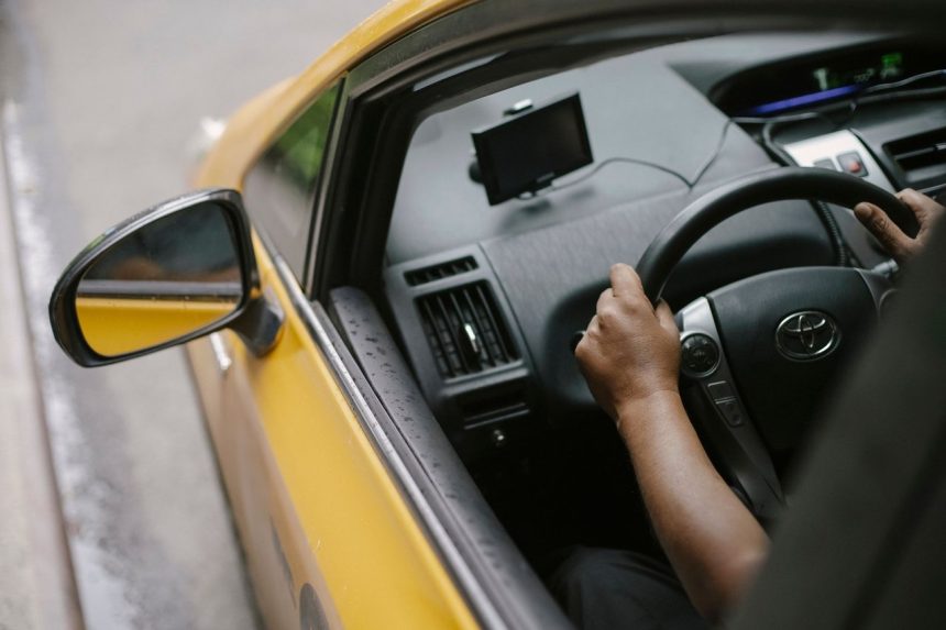 Vista superior de uma pessoa dirigindo um táxi amarelo equipado com navegador GPS em uma rua urbana durante o dia.