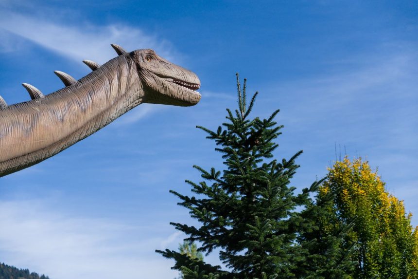 Escultura em tamanho real de um dinossauro saurópode ao ar livre, diante de árvores e do céu azul.